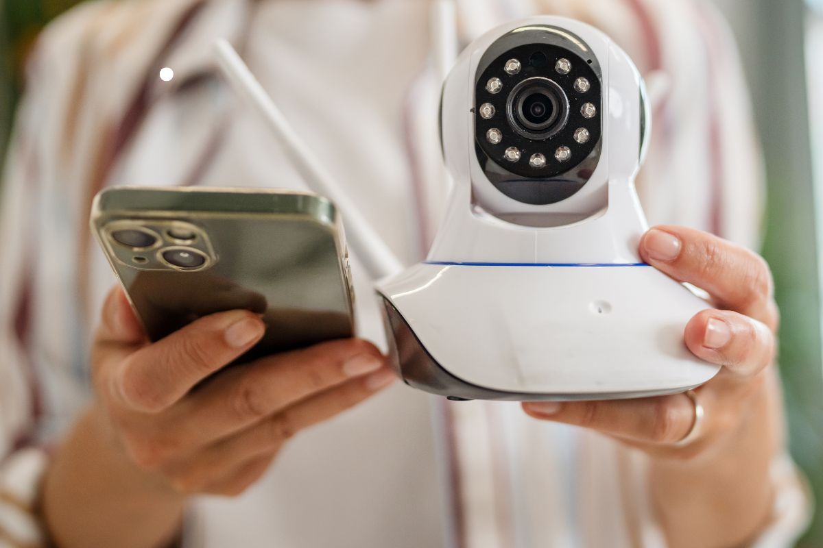 A person holds a smartphone in one hand and a security camera in the other, highlighting modern home monitoring technology.