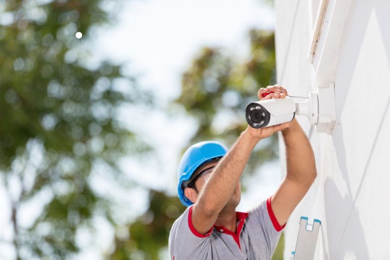 A worker in a blue helmet installs a security camera on a white wall, surrounded by greenery on a sunny day.