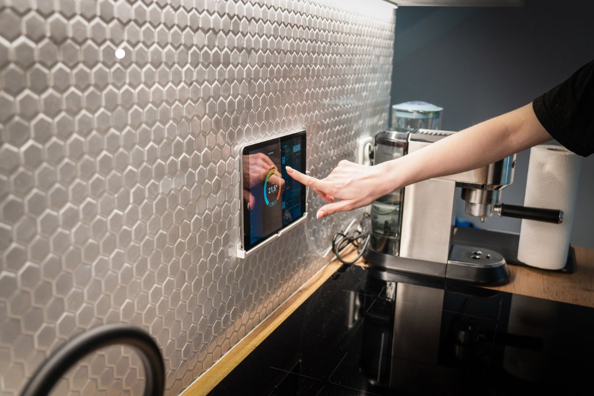 A hand touches a touchscreen panel embedded in a modern kitchen wall, displaying a temperature reading amid sleek appliances.