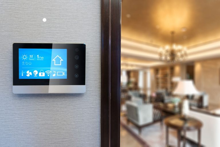 A modern touchscreen control panel displaying weather, time, and home settings, mounted on a textured wall with a stylish living room blurred behind.