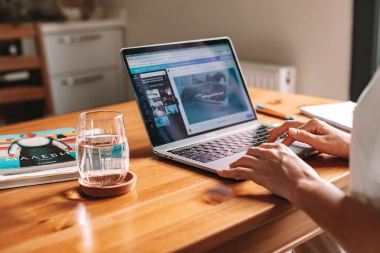 A person typing on a laptop with a dog video displayed, beside a glass of water and an art book on a wooden table.