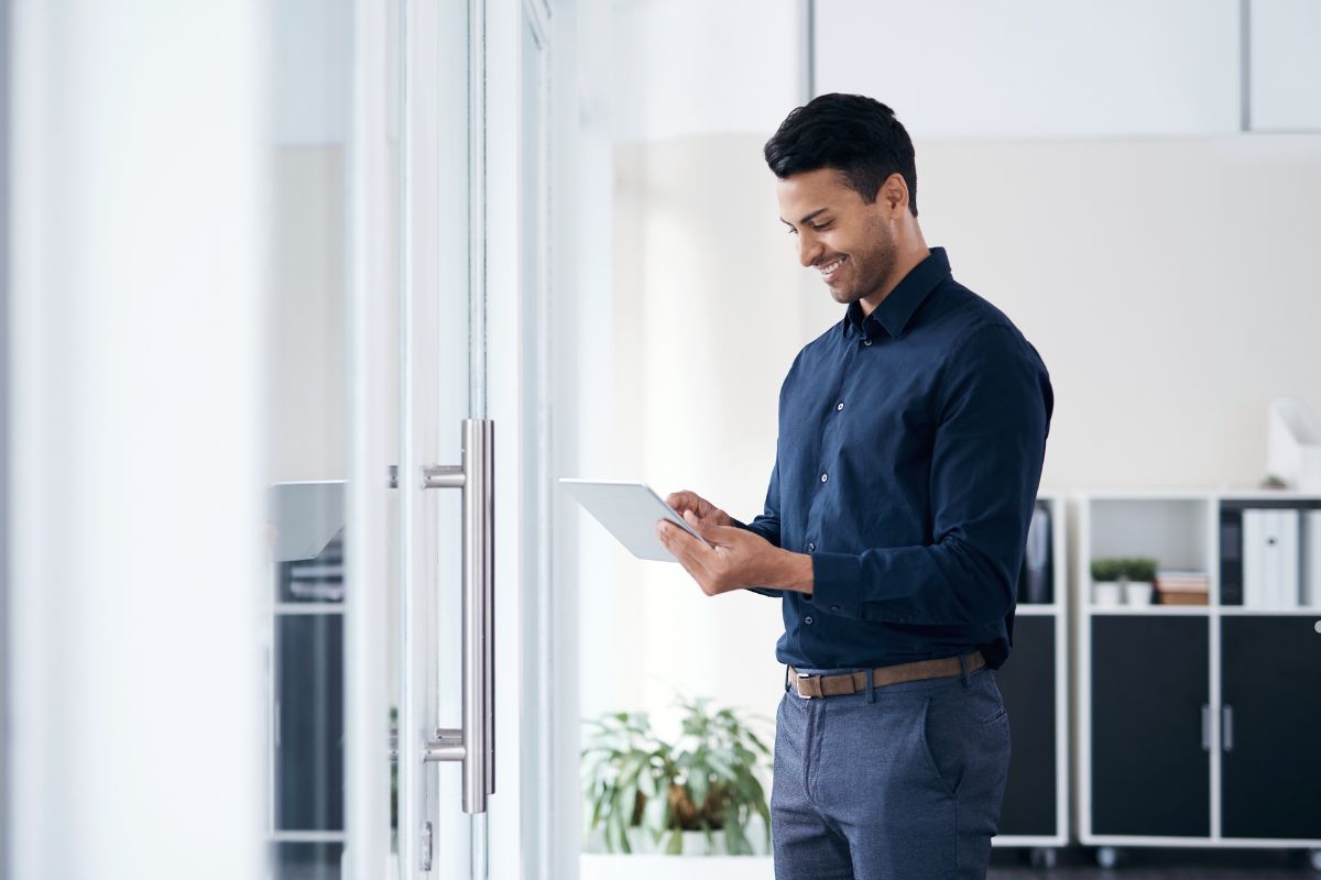A man in a navy shirt stands by a glass door, holding a tablet and looking thoughtfully at the screen in a modern office setting.